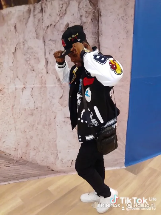 Young Man in Varsity Jacket Struts Confidently in Front of a Stone Wall Background – AI Generated Video | Hailuo AI